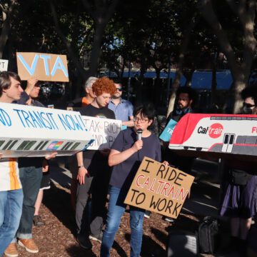 Photo of about 30 people standing outside a building. Nearly everyone is holding signs with slogans such as "Fund transit now" and "Support Public Transit." A shorter woman with short black hair and glasses is holding a sign reading "I ride Caltrain to work" and is speaking into a microphone
