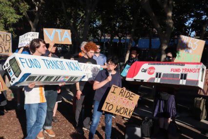 Photo of about 30 people standing outside a building. Nearly everyone is holding signs with slogans such as "Fund transit now" and "Support Public Transit." A shorter woman with short black hair and glasses is holding a sign reading "I ride Caltrain to work" and is speaking into a microphone