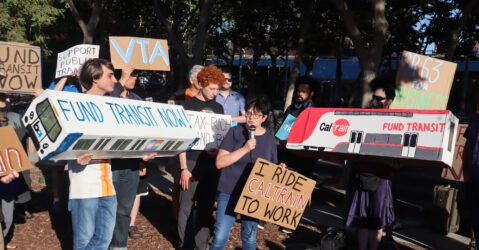 Foto de unas 30 personas afuera de un edificio. Casi todas portan carteles con lemas como "Financien el transporte público ahora" y "Apoyen el transporte público". Una mujer de baja estatura, con cabello negro corto y gafas, sostiene un cartel que dice "Voy en Caltrain al trabajo" y habla por un micrófono.
