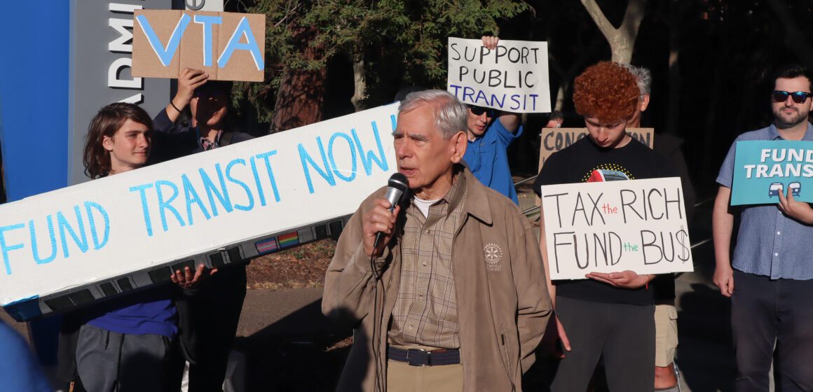 Photo of a crowd of people standing outside, holding signs reading "Fund transit now" and "Tax the rich, fund the bus" and "Support public transit." The photo is centered on Rod Diridon, an elderly man with white hair wearing a beige jacket with the Rotary Club logo
