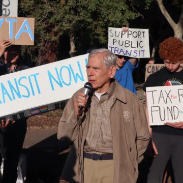 Photo of a crowd of people standing outside, holding signs reading "Fund transit now" and "Tax the rich, fund the bus" and "Support public transit." The photo is centered on Rod Diridon, an elderly man with white hair wearing a beige jacket with the Rotary Club logo