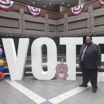 Một người đàn ông mặc vest đứng trước những chữ cái lớn có dòng chữ VOTE