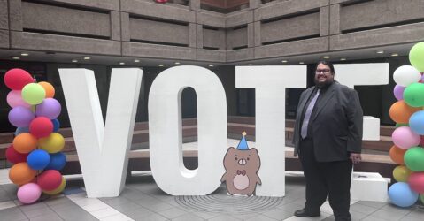 A man in a suit stands in front of large letters reading VOTE