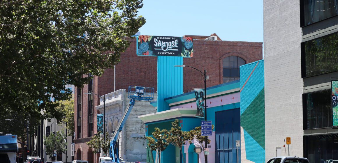 Photo of Santa Clara Street, focused on the blue and green facade of the vacant former Chase Bank. A large sign coming off the building reads "Welcome to San Jose Downtown", visible to the cars driving along the street below