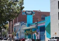 Photo of Santa Clara Street, focused on the blue and green facade of the vacant former Chase Bank. A large sign coming off the building reads "Welcome to San Jose Downtown", visible to the cars driving along the street below