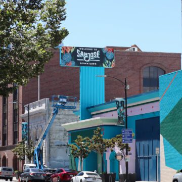 Photo of Santa Clara Street, focused on the blue and green facade of the vacant former Chase Bank. A large sign coming off the building reads "Welcome to San Jose Downtown", visible to the cars driving along the street below