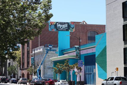 Photo of Santa Clara Street, focused on the blue and green facade of the vacant former Chase Bank. A large sign coming off the building reads "Welcome to San Jose Downtown", visible to the cars driving along the street below