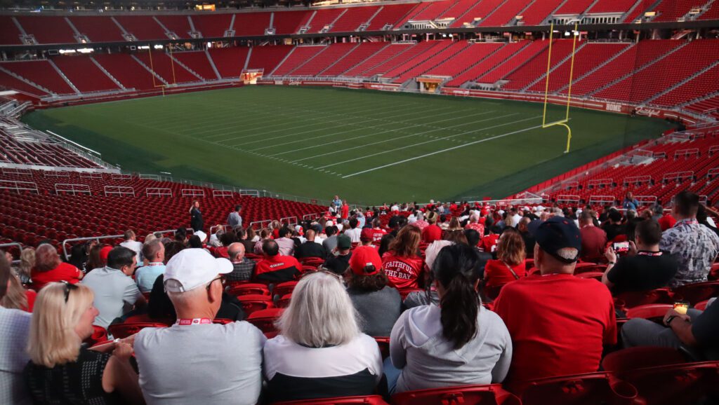 Photo of dozens of people wearing red, white, and gold, sitting in one section of Levi's Stadium, with the empty field and other empty sections visible in the background