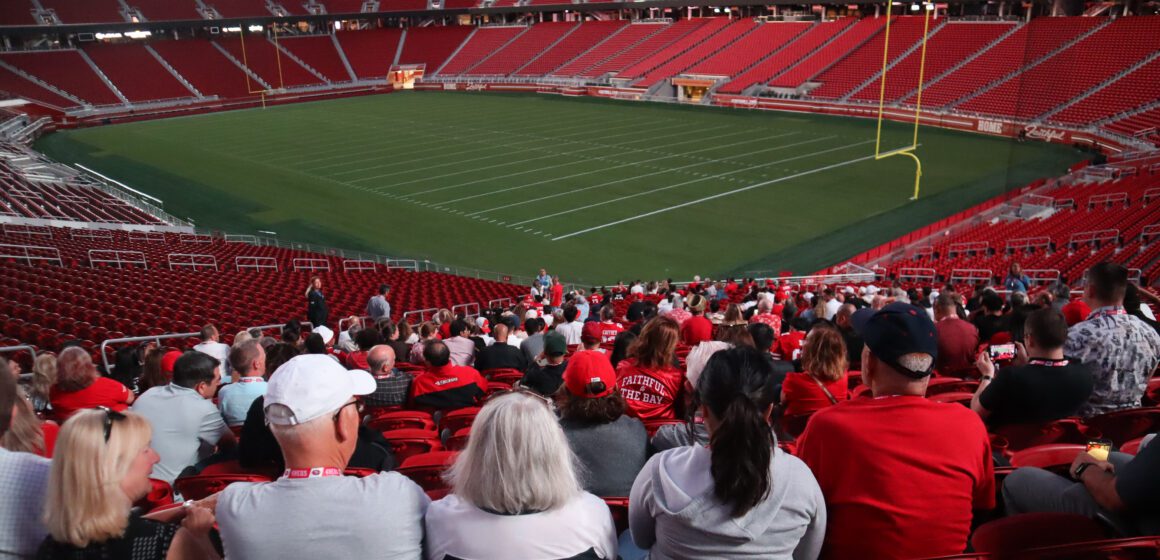 Photo of dozens of people wearing red, white, and gold, sitting in one section of Levi's Stadium, with the empty field and other empty sections visible in the background