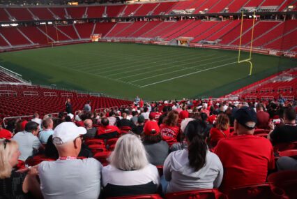 Photo of dozens of people wearing red, white, and gold, sitting in one section of Levi's Stadium, with the empty field and other empty sections visible in the background