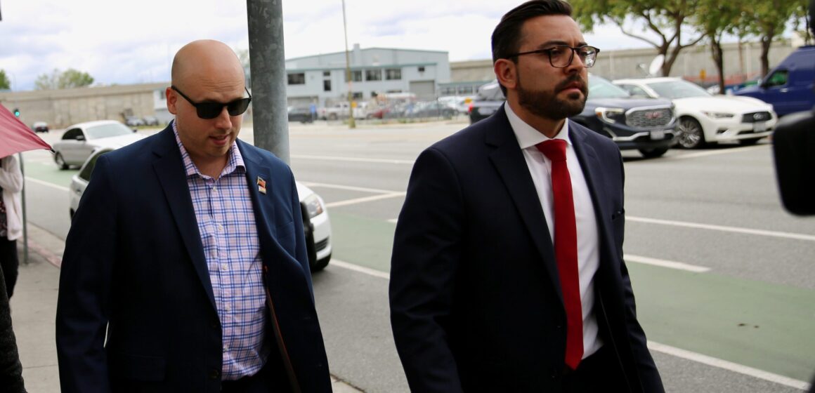 A man in a blazer walks on the sidewalk next to a man in a suit and tie