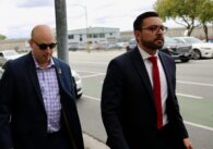 A man in a blazer walks on the sidewalk next to a man in a suit and tie