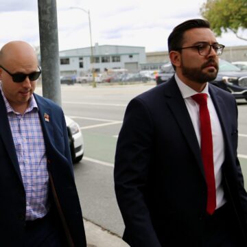A man in a blazer walks on the sidewalk next to a man in a suit and tie