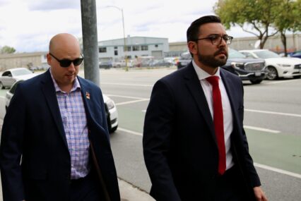 A man in a blazer walks on the sidewalk next to a man in a suit and tie