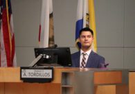 A man in a suit and tie sits behind a computer at a dais