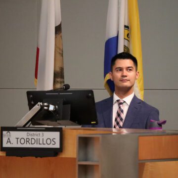 A man in a suit and tie sits behind a computer at a dais