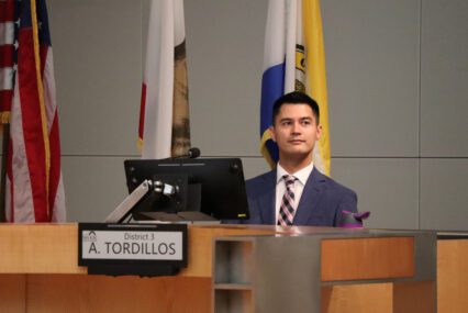 A man in a suit and tie sits behind a computer at a dais