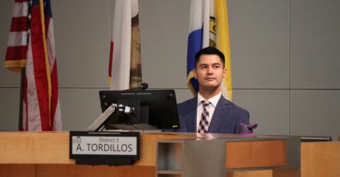 A man in a suit and tie sits behind a computer at a dais
