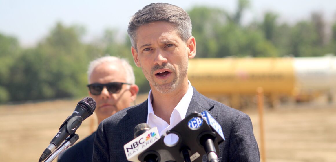 A man in a suit jacket speaks into microphones at a podium during an outdoor press conference