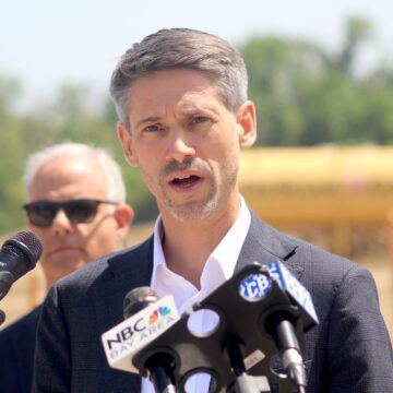 A man in a suit jacket speaks into microphones at a podium during an outdoor press conference