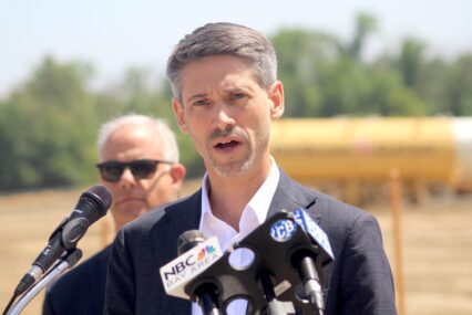 A man in a suit jacket speaks into microphones at a podium during an outdoor press conference