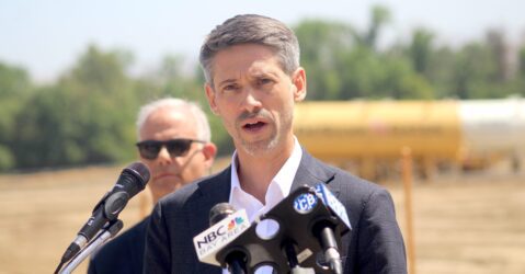 A man in a suit jacket speaks into microphones at a podium during an outdoor press conference