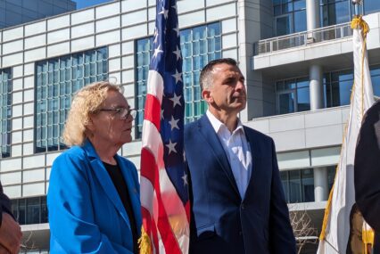 An older woman and middle-aged man stand next to an American flag