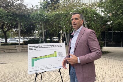 A U.S. congressman in a suit and tie speaks outside next to a chart about federal funding cuts