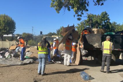 People in reflective vests throw trash from a homeless encampment into a dumpster