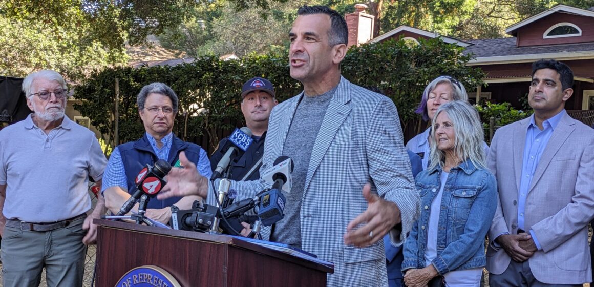 A man in a blazer speaks at a podium, with a line of people behind him