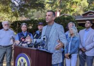 A man in a blazer speaks at a podium, with a line of people behind him