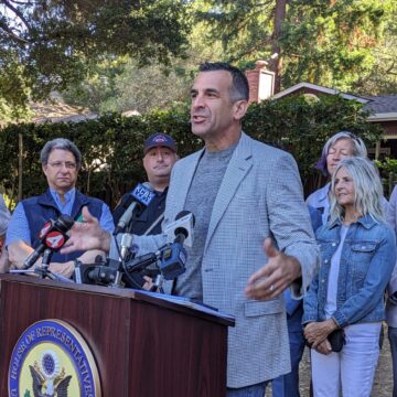 A man in a blazer speaks at a podium, with a line of people behind him