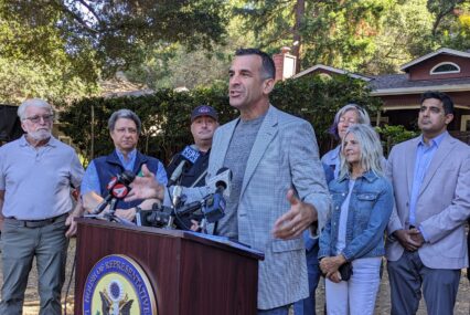 A man in a blazer speaks at a podium, with a line of people behind him