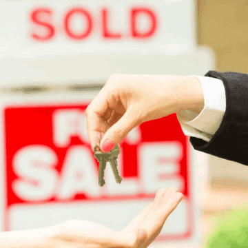 A hand passes house keys to another hand with a for sale sign in the background