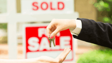 A hand passes house keys to another hand with a for sale sign in the background