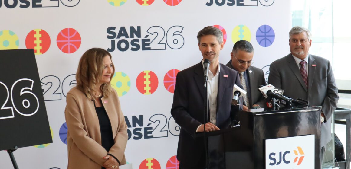 Photo of four people, with one man in a black suit coat and white button down shirt speaking at a podium labeled "SJC San Jose Mineta International" in front of a backdrop with sporty icons and the label "San Jose 26"