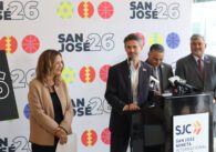 Photo of four people, with one man in a black suit coat and white button down shirt speaking at a podium labeled "SJC San Jose Mineta International" in front of a backdrop with sporty icons and the label "San Jose 26"