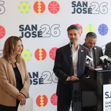 Photo of four people, with one man in a black suit coat and white button down shirt speaking at a podium labeled "SJC San Jose Mineta International" in front of a backdrop with sporty icons and the label "San Jose 26"
