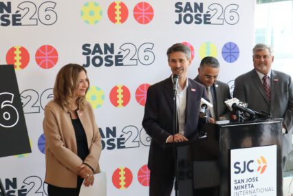 Photo of four people, with one man in a black suit coat and white button down shirt speaking at a podium labeled "SJC San Jose Mineta International" in front of a backdrop with sporty icons and the label "San Jose 26"