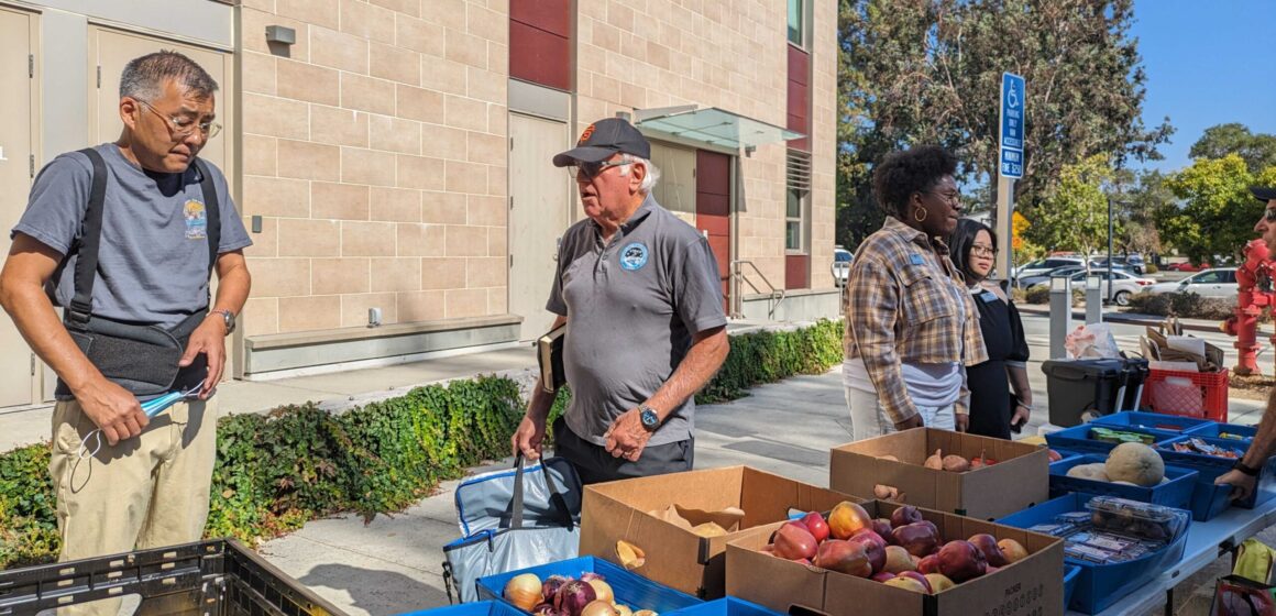 People stand behind tables with boxes of food, including onions and apples