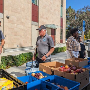 People stand behind tables with boxes of food, including onions and apples