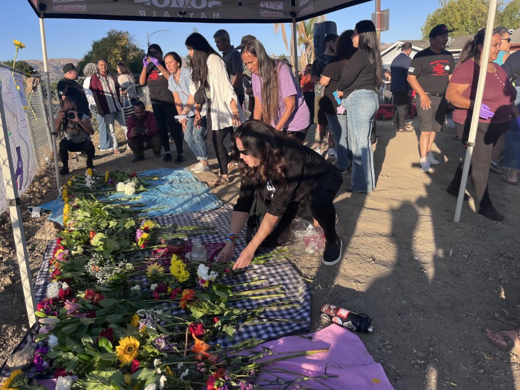 A woman bends down to place flowers on the ground, with a crowd of people standing behind her