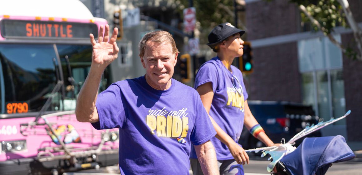 Foto de un hombre mayor de cabello rubio y blanco saludando y sonriendo a la cámara, con una camiseta morada que dice "Viaja con Orgullo". Detrás de él, una persona negra más alta, con la misma camiseta y una pulsera de arcoíris, empuja un cochecito. Detrás de ambos, un autobús rosa brillante desenfocado.