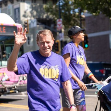 Photo of an older man with white and blonde hair waving and smiling at the camera, wearing a purple shirt that read "ride with Pride." Behind him is a taller Black person wearing the same shirt and a rainbow wristband pushing a stroller. Behind both of them is an out-of-focus bright pink bus.