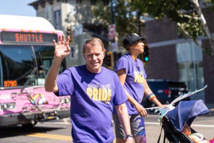 Photo of an older man with white and blonde hair waving and smiling at the camera, wearing a purple shirt that read "ride with Pride." Behind him is a taller Black person wearing the same shirt and a rainbow wristband pushing a stroller. Behind both of them is an out-of-focus bright pink bus.