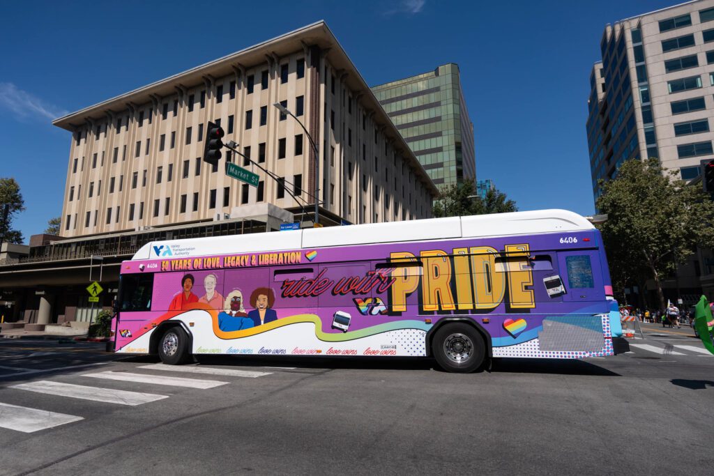 Foto del Autobús del Orgullo de la VTA circulando por la intersección de las calles Santa Clara y Market en el centro de San José. El autobús presenta un degradado de rosa a morado y retratos dibujados de cuatro líderes de la comunidad LGBTQ+, entre ellos Ken Yeager y Billy DeFrank. El autobús dice "50 años de Amor, Legado y Liberación" y "Viaja con Orgullo".