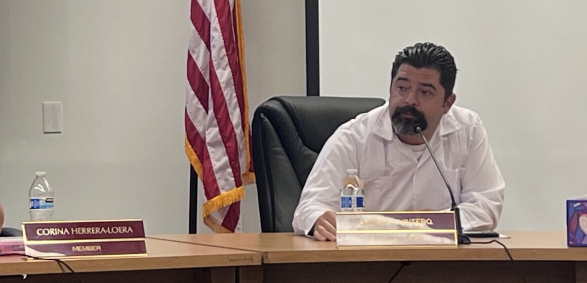A man in a white shirt sits at a desk during a school board meeting, with an American flag behind him