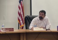 A man in a white shirt sits at a desk during a school board meeting, with an American flag behind him