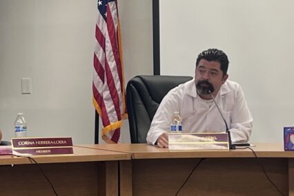 A man in a white shirt sits at a desk during a school board meeting, with an American flag behind him