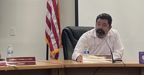 A man in a white shirt sits at a desk during a school board meeting, with an American flag behind him